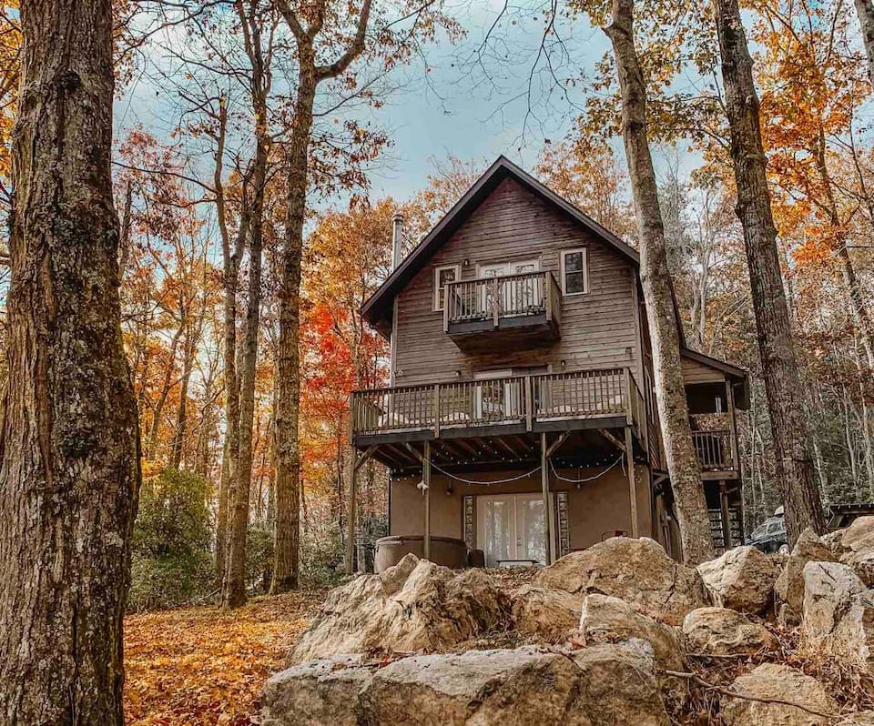 Beautiful multilevel brown wooden cabin surrounded with autumn leaves on trees with large rocks in foreground.