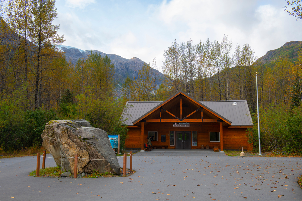 big rock with fencing placed in front of a wooden building with roof surrounding by trees and mountains in the backdrop