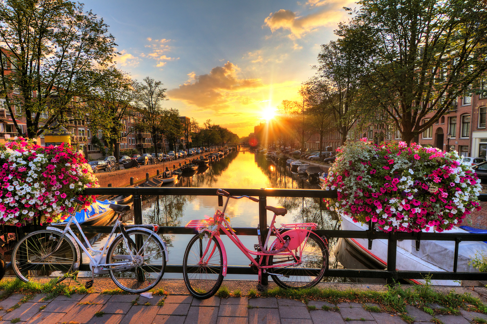 sun peeking from behind the buildings over a river channel with a bridge with bikes on it traveling to amsterdam
