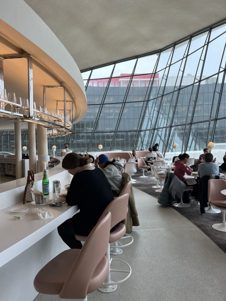 Inside the cafe at the TWA hotel at JFK. There are people sat at tables with huge glass windows behind them. 