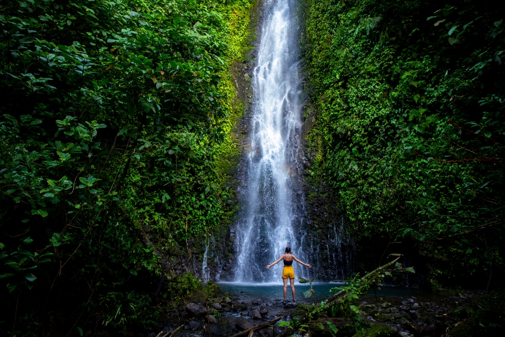 Girl stands in front of a mighty waterfall with her hands raised in the air, Article is about travelling to Costa Rica