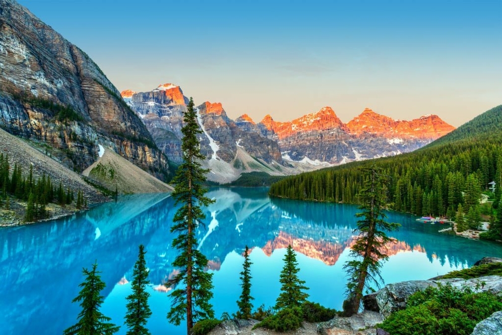 sunrise over mountains whose reflection can be seen in a blue lake surrounded by green trees icefields parkway