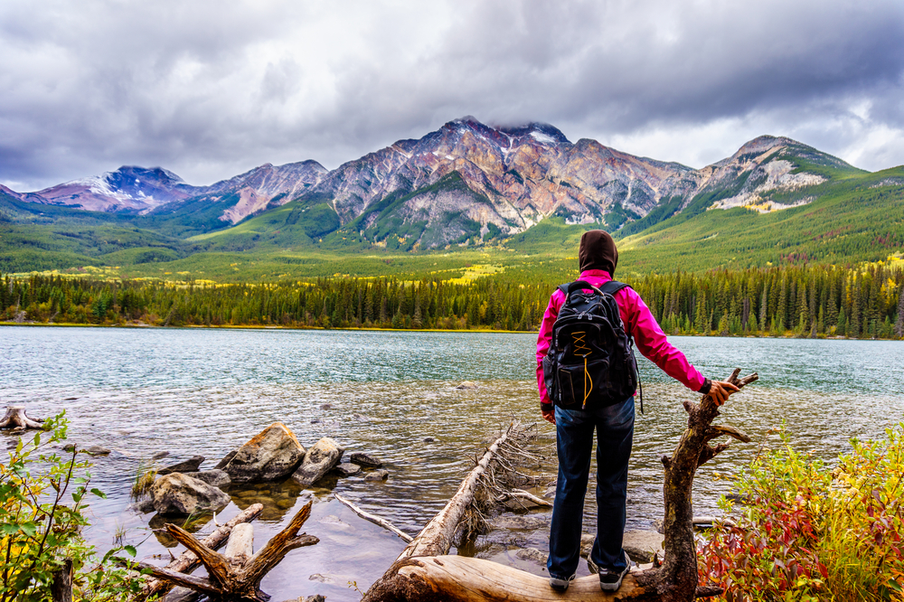 woman hiking and standing at the edge of a lake with trees and mountains in front
