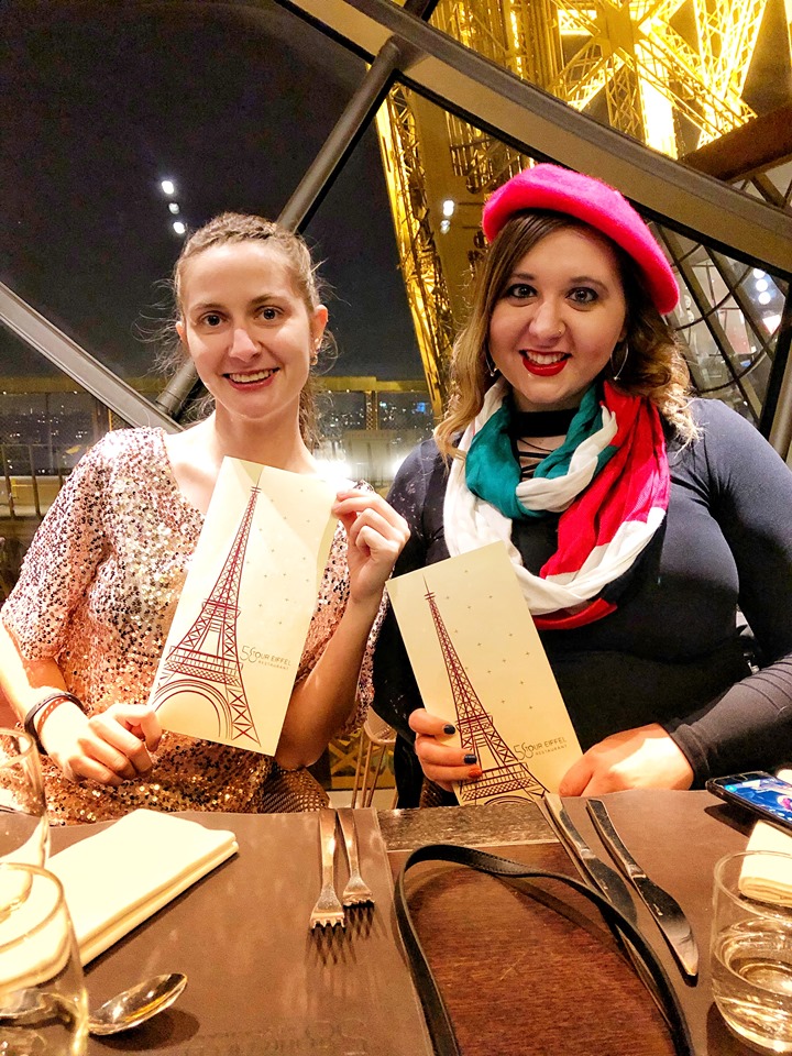 My two young daughters holding menus at a table in the 58 Tour Eiffel restaurant with Eiffel Tower views out the window behind them.