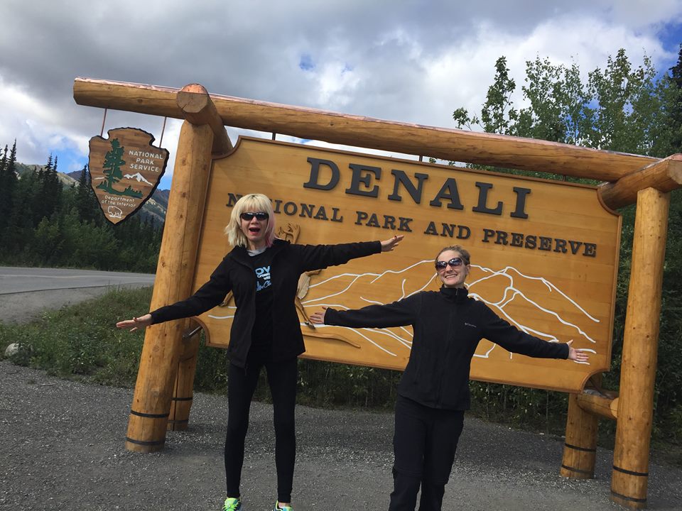 Two Caucasian women with smiles stand in front of wooden Denali welcome sign on an Alaska itinerary.