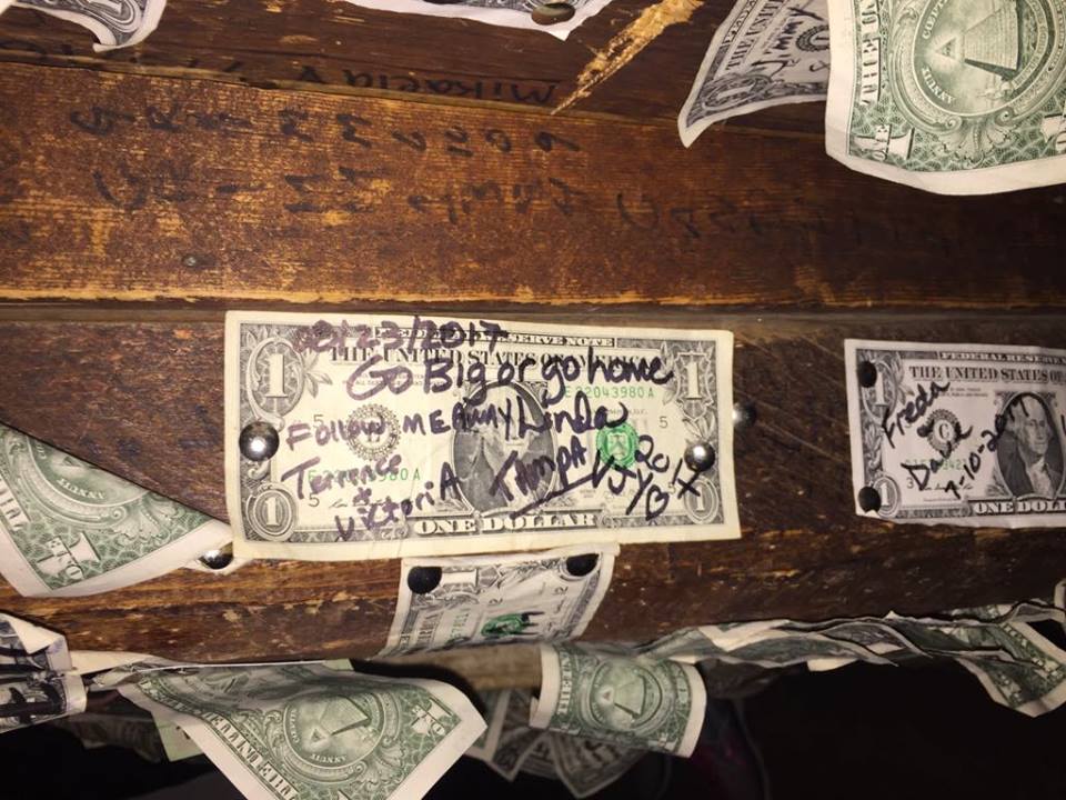 A dollar bill tacked to a wooden wall signed in black by me, my daughter, and her boyfriend with other dollars around it.