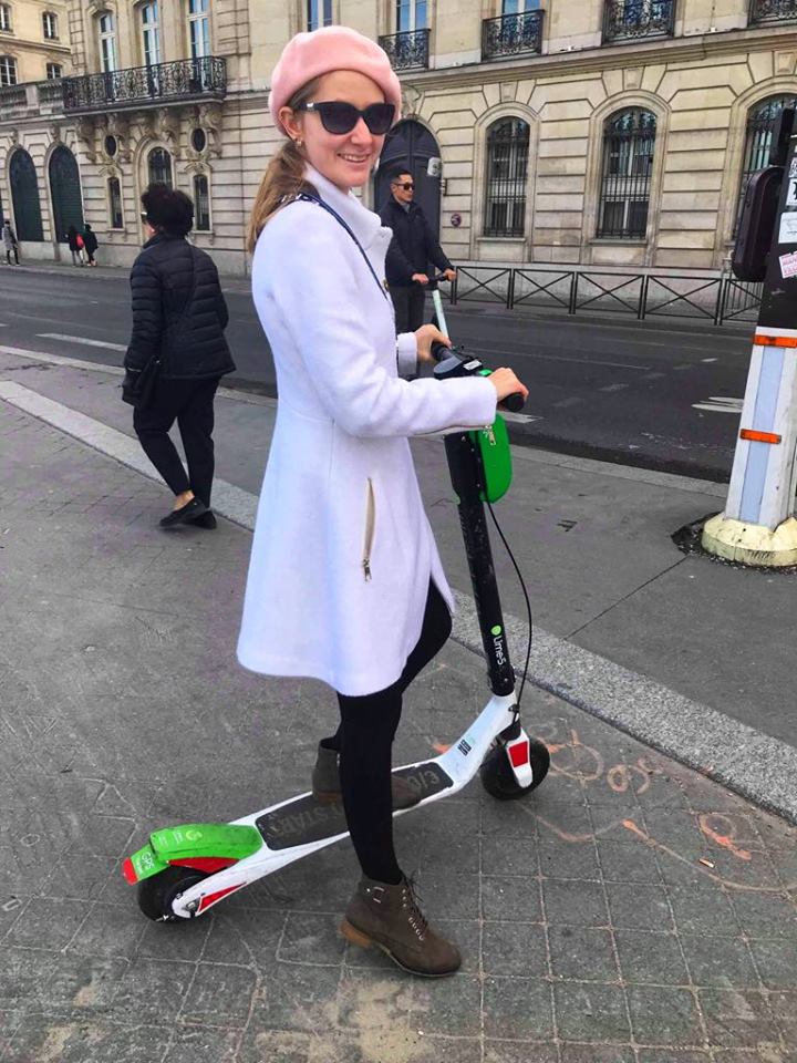 Women in a pink beret and a white coast on a scoter on a road in Paris. 