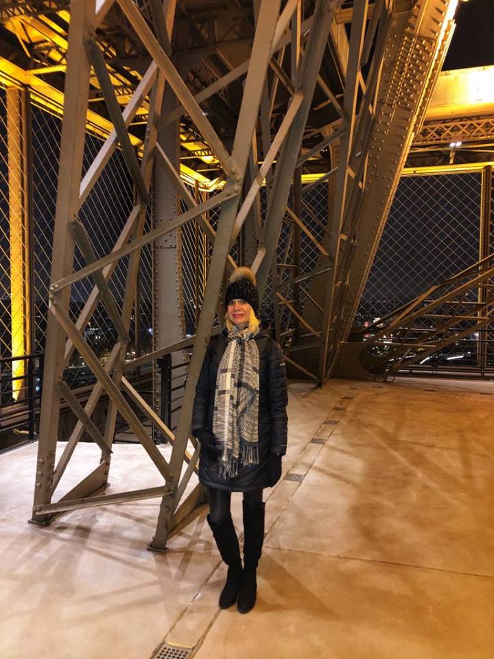 Linda stood in the Eiffel Tower. She is wearing a winter coat scarf and bobble hat.  