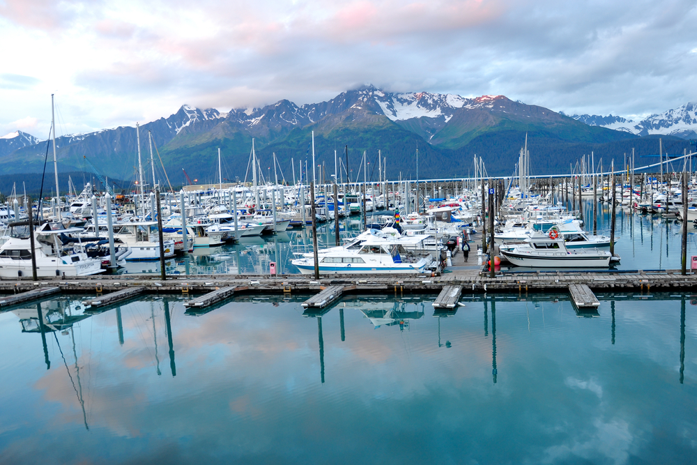 Seward Alaska marina with boats in slips. Snow capped mountains in background and blue water in foreground.