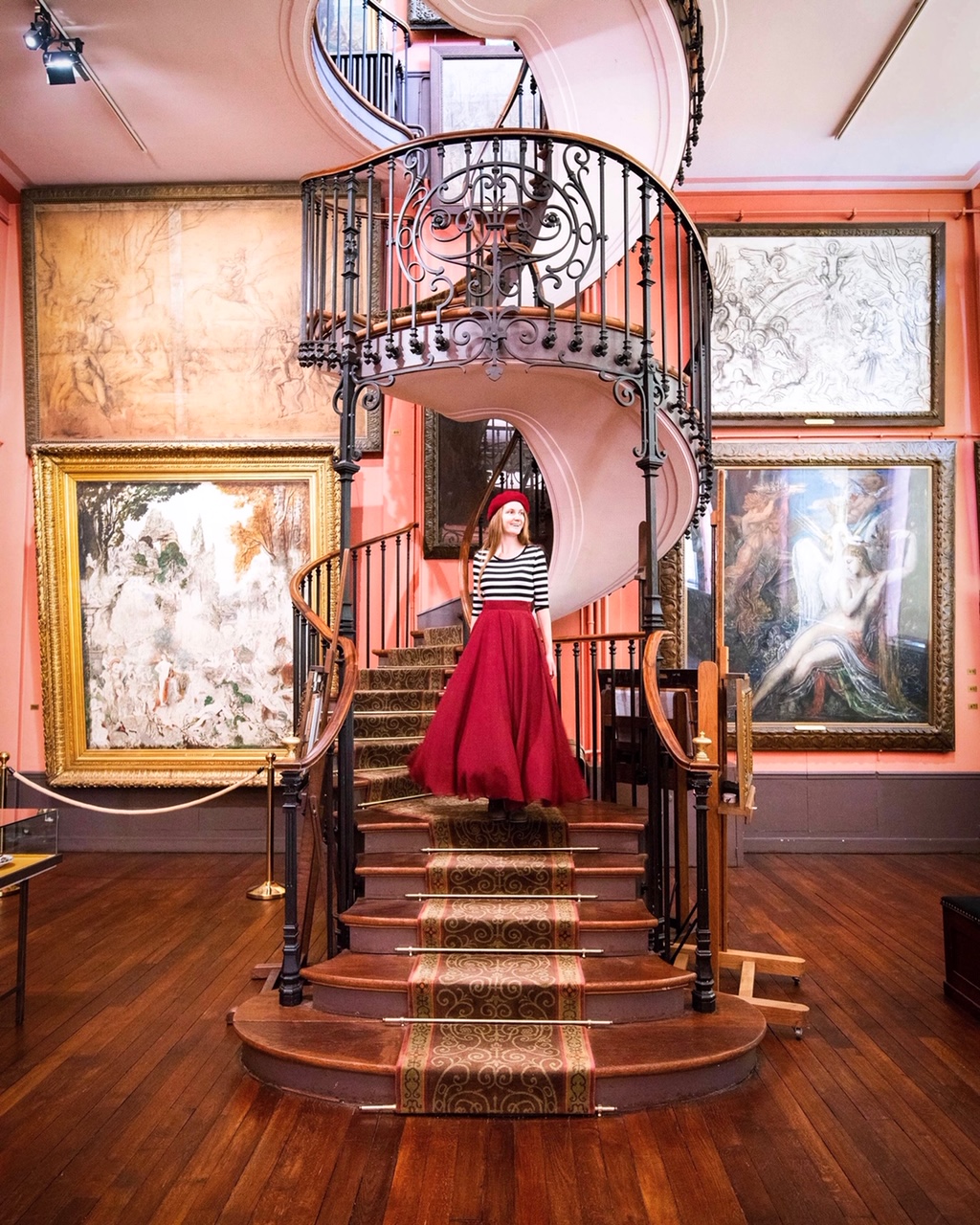 Young woman in a red shirt and matching beret on spiral staircase in Gustave Moreau Museum with paintings on the wall.