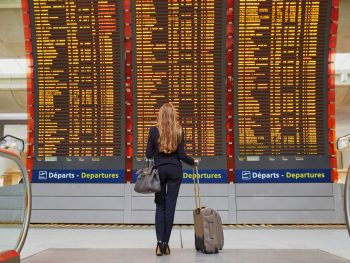 Female with blonde hair looks over flight information screen while holding onto her carry-on suitcase and matching gray bag.