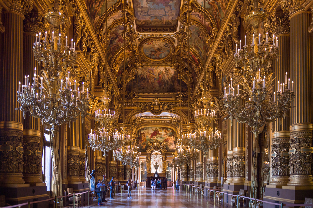 Extravagant gold room with opulent chandeliers lining both sides. People looking at paintings on walls.