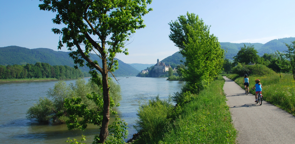 People riding bikes on a flat, gravel path long the tree-lined river with rolling hills and Passau in the distance.