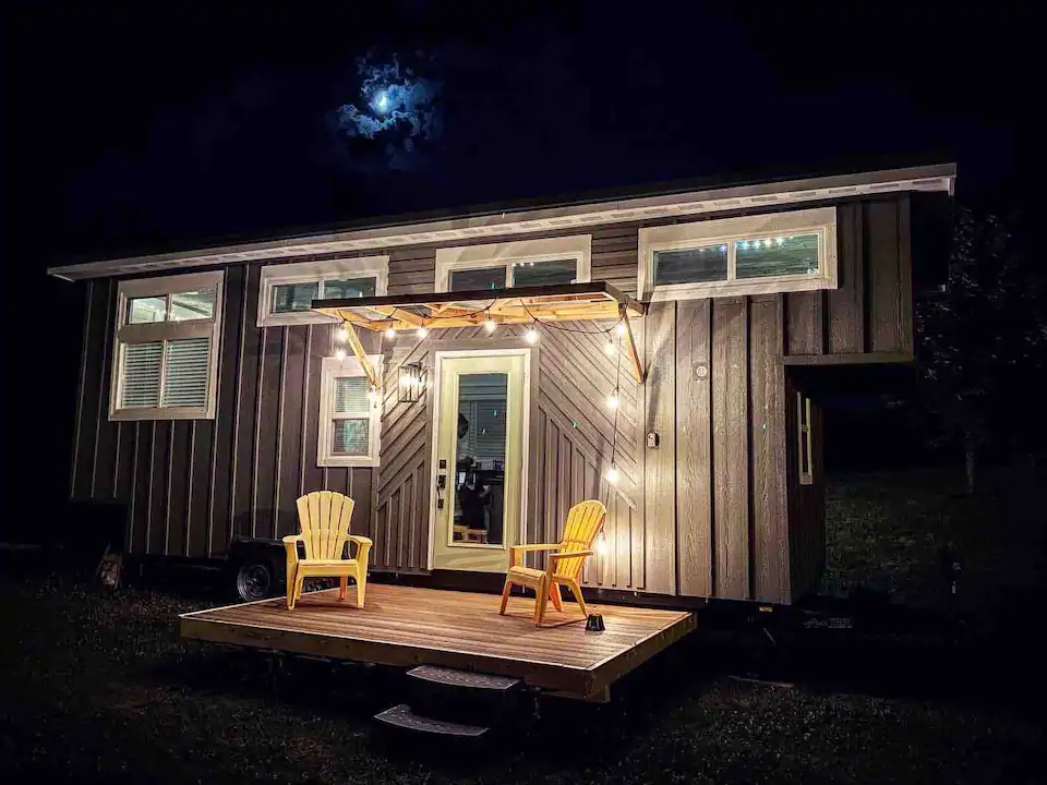 Photo of cute tiny house with 2 chairs on deck, and lights illuminating the front entrance.