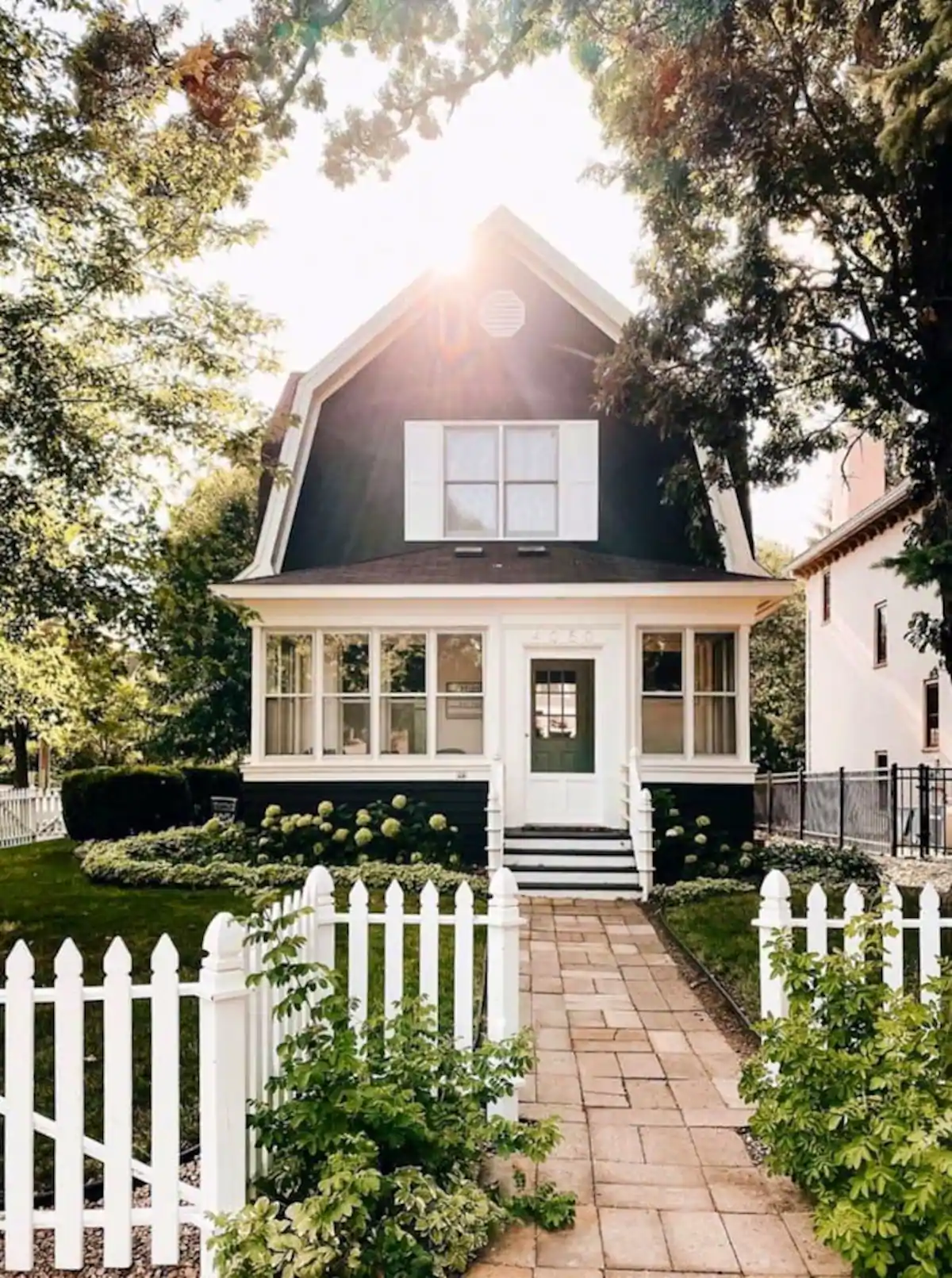 Sun peeking over the roof of a charming Airbnb in Minneapolis with dark exterior, white picket fence in front of it with charming walkway.