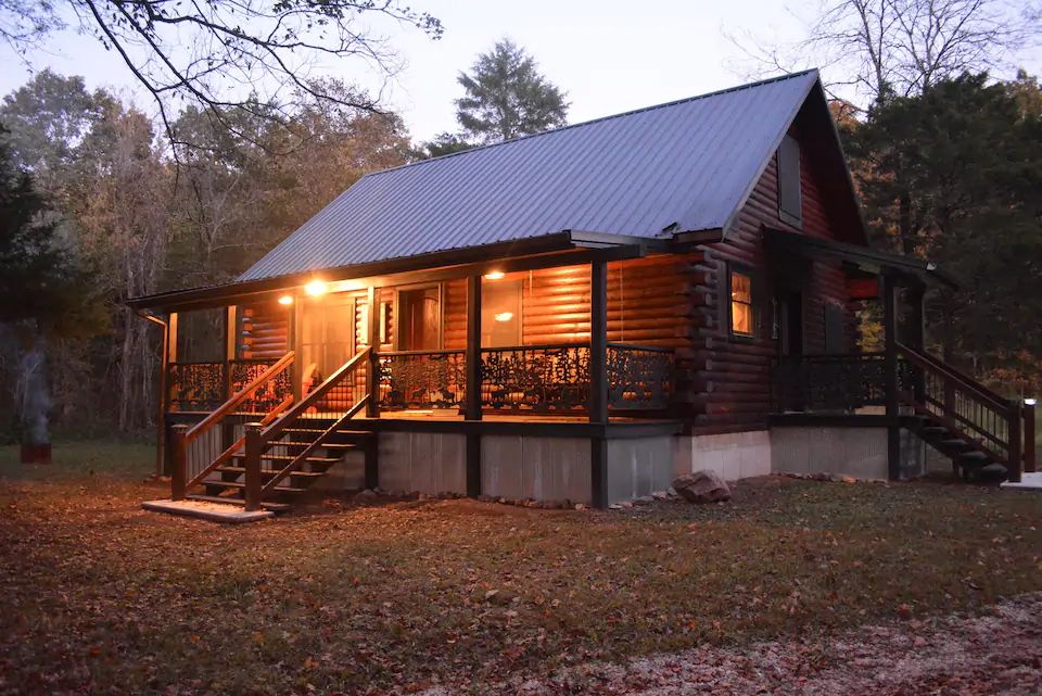 Charming cabin at dusk brightly lit. Front porch has chairs to sit in.