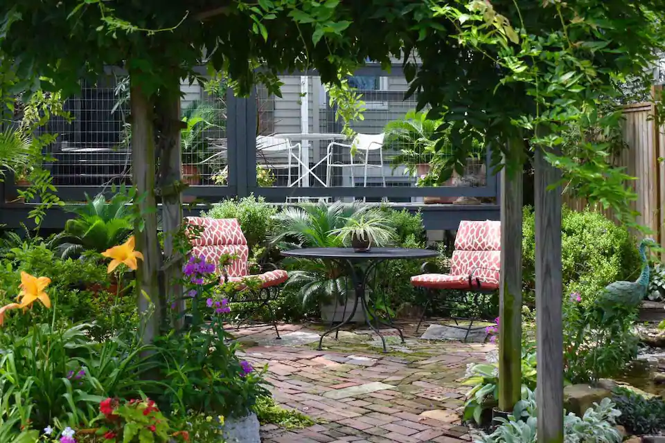 Photo of lush garden with red brick walk, and flowers blooming. Two red chairs are around a black table.