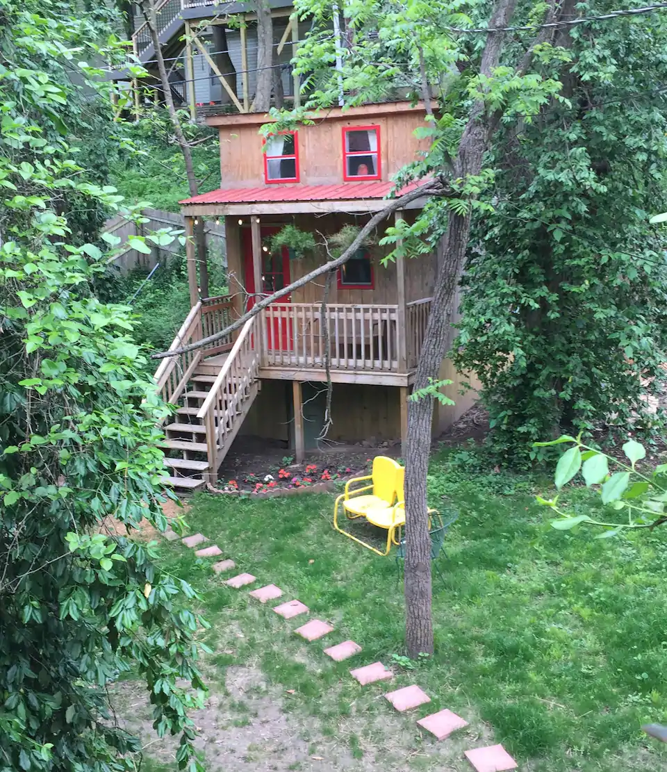 A photo of a cute tiny house surrounded by trees, It has red trim around the windows and yellow chairs in the yard.