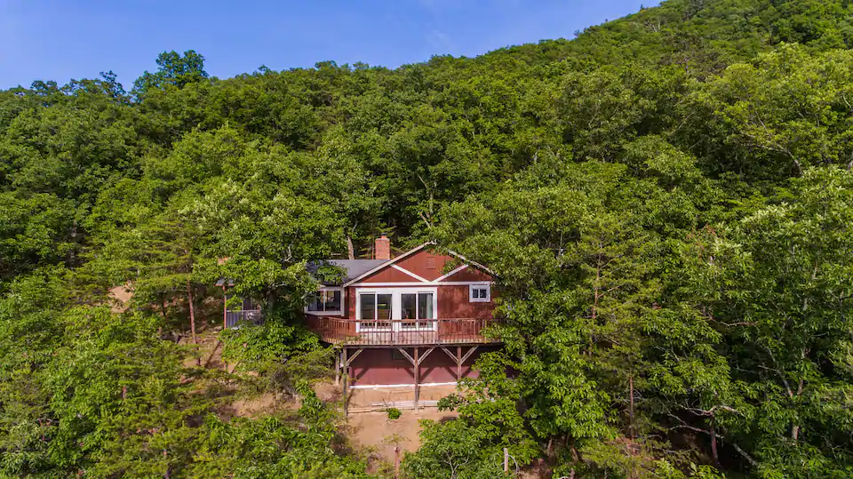 Photo of red West Virginia Airbnb cabin perched on side of mountain surrounded by lush green trees.
