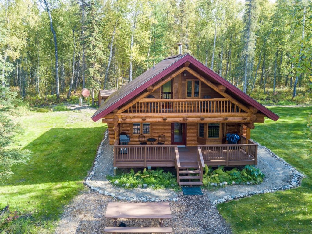 One of the cozy log cabins in Alaska this one has a red roof and generous front porch situated in a large green yard.