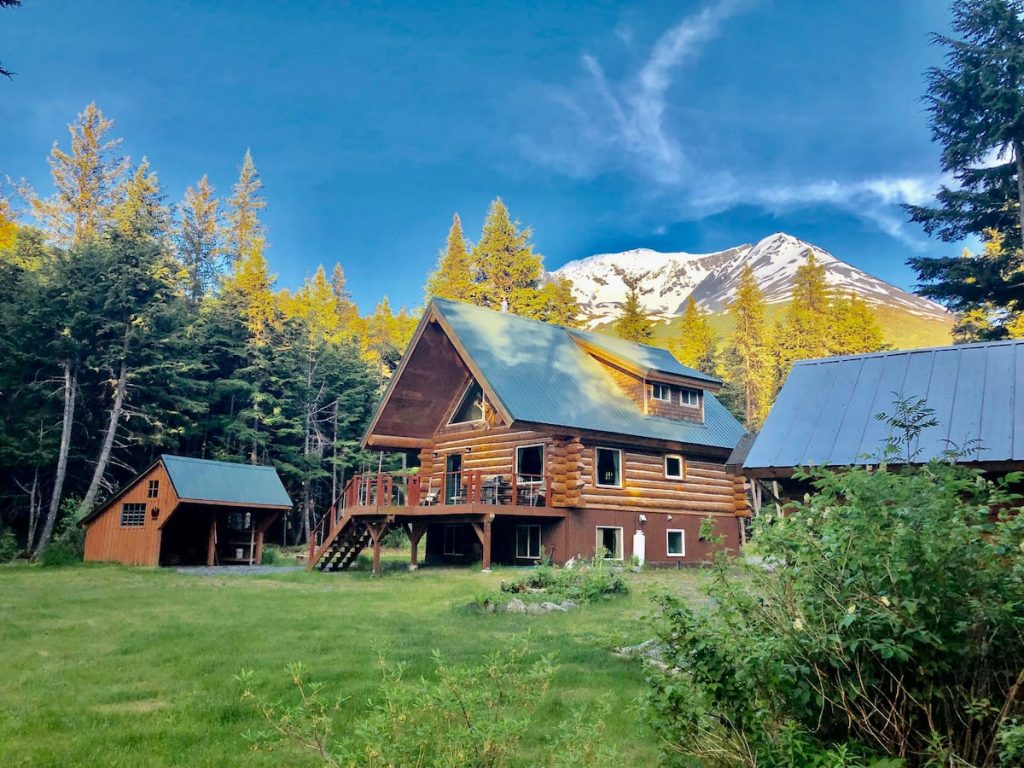 This is one of the best log cabins in Alaska and this photo shows it nestled in the snow-capped mountains. Cabin has green metal roof and large windows,