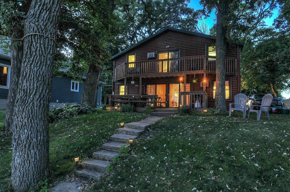 Photo of large 2 story cabin with warm lights from indoors lighting it. There are 2 chairs, and steps leading down through a green yard.