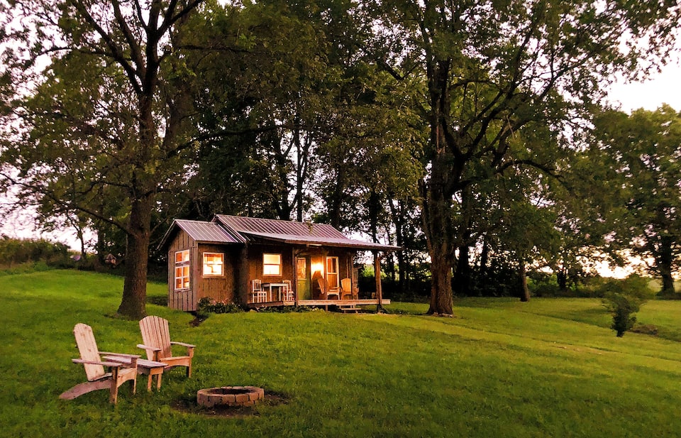 Rustic cabin set on hillside with bright lights shining inside surrounded by tall green trees, with Adirondack chairs surrounding firepit.