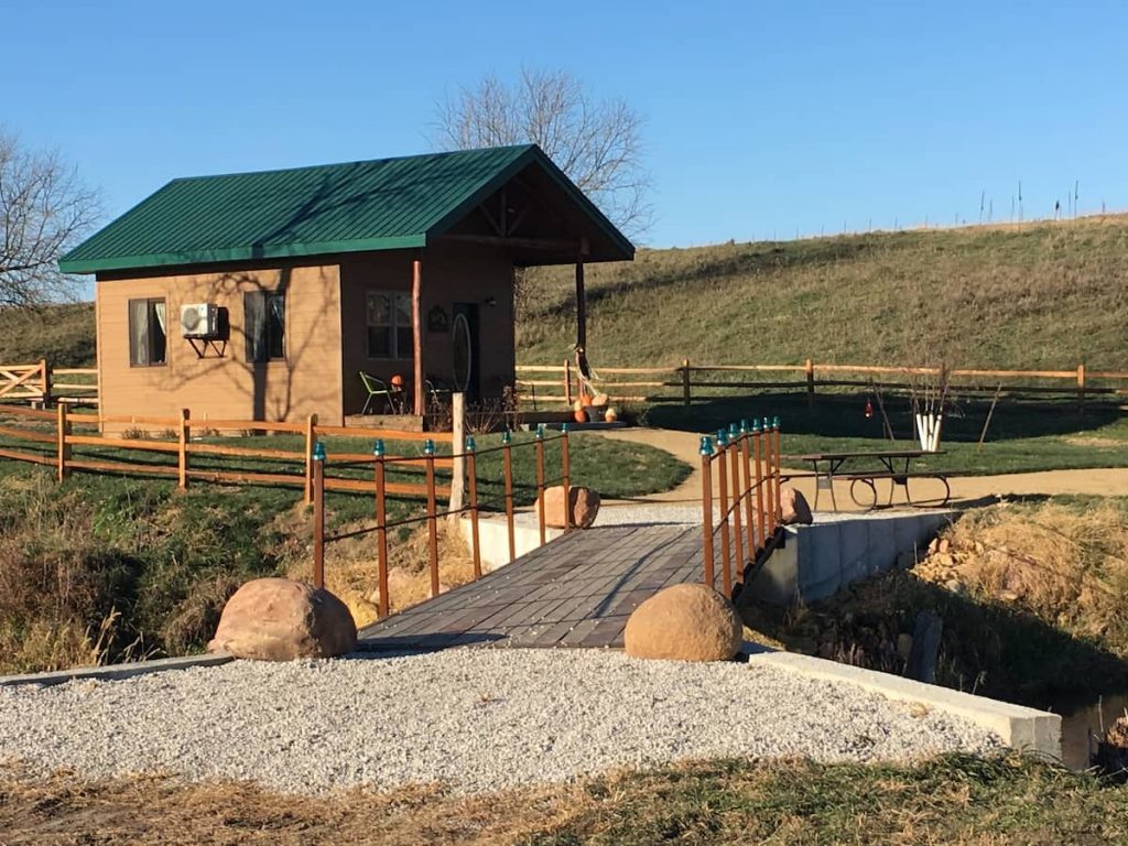 Log cabin with green roof and chairs on front porch. Bridge to cabin in foreground.