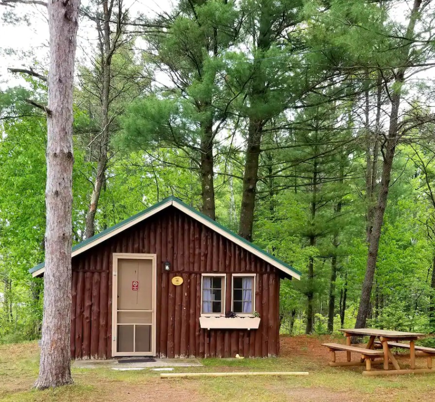 The exterior of a classic log cabin with two windows and a window flower box. Next to it is a picnic table and it is surrounded by trees.
