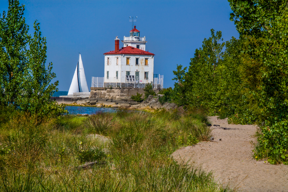 A sandy beach that also has grasses and trees near a large lighthouse that has a sail boat next to it
