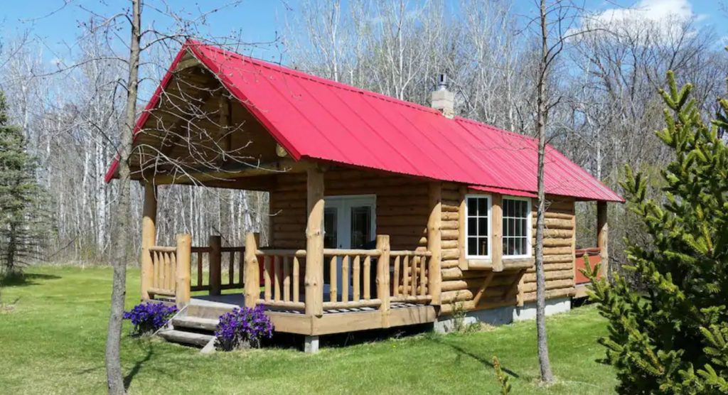 The exterior of a one room classic log cabin with a red roof and a large covered front porch