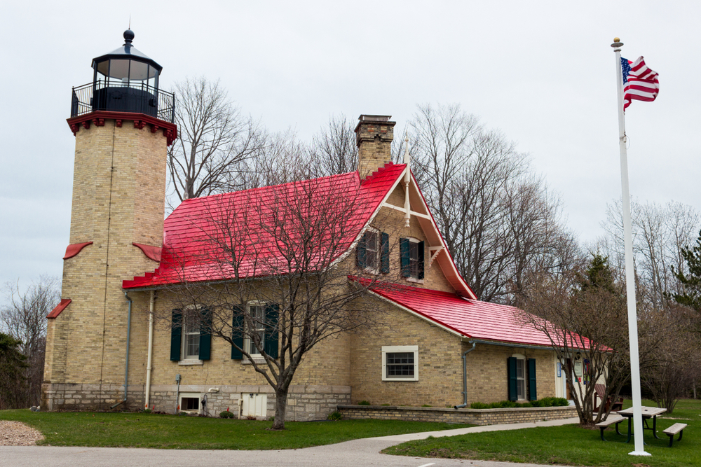 A tan brick lighthouse with a red roof and surrounded by a green lawn and trees in the middle of winter