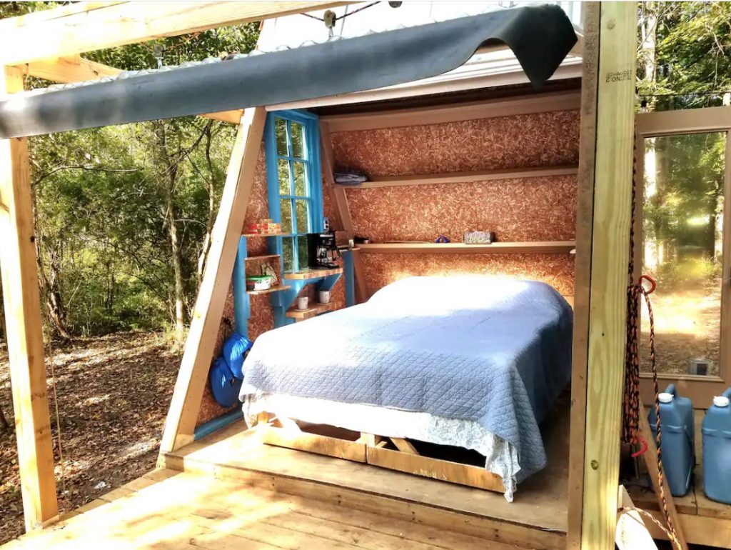 The inside view of the Tiny A-frame with the side of the building lifted, so you can see the queen sized bed and small shelving areas