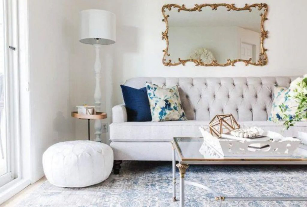 A luxuriously decorated living room with a gold gilt mirror, a lage sofa, and a glass coffee tabl