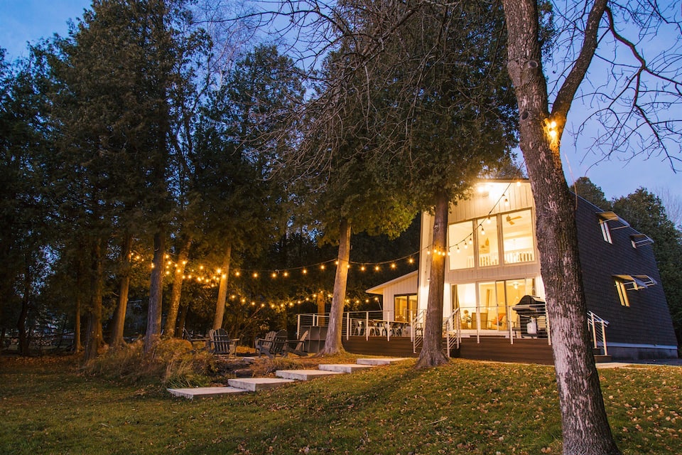 Twilight photo of a Door County cabin with floor to ceiling windows, large deck and outdoor white lights illuminating the back yard.