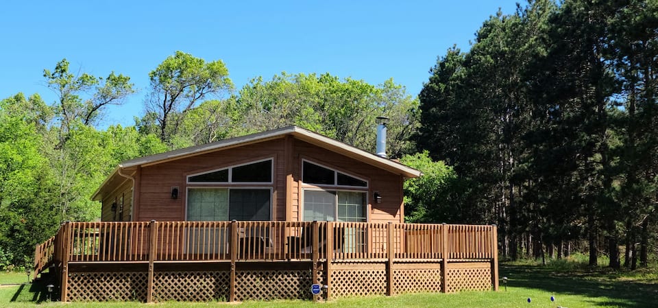 Cozy cabin In Wisconsin with wooden fence around wraparound porch, surrounded by large green yard.