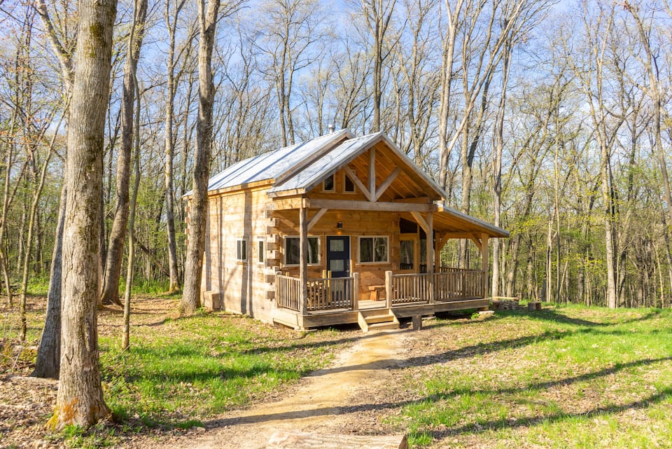 Lovely log cabin in Wisconsin with cute front porch, surrounded by a forest of trees with sun brightly shining.