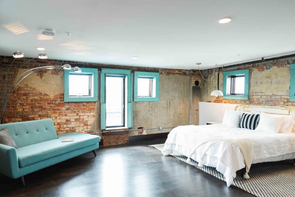 A large bedroom with exposed brick walls, a cozy looking bed, and a modern sofa, with the window trims painted teal