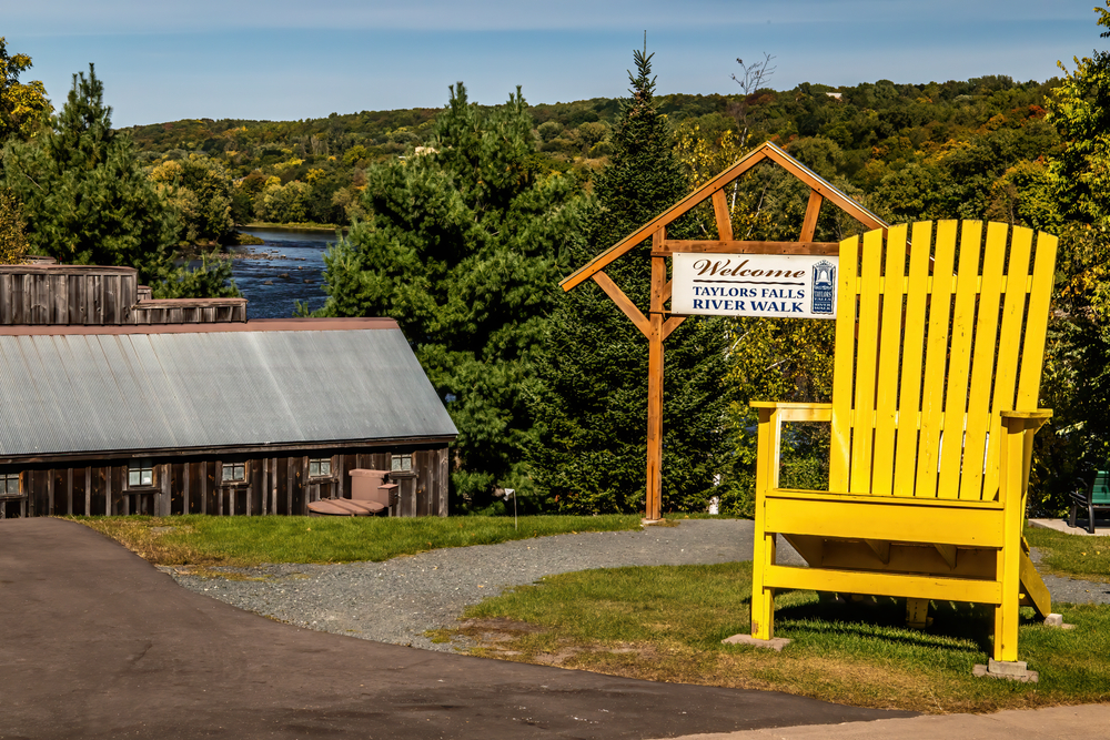 the entrance to the Taylors Falls River Walk that has a massive yellow Adirondack chair in front of it with the river between the trees