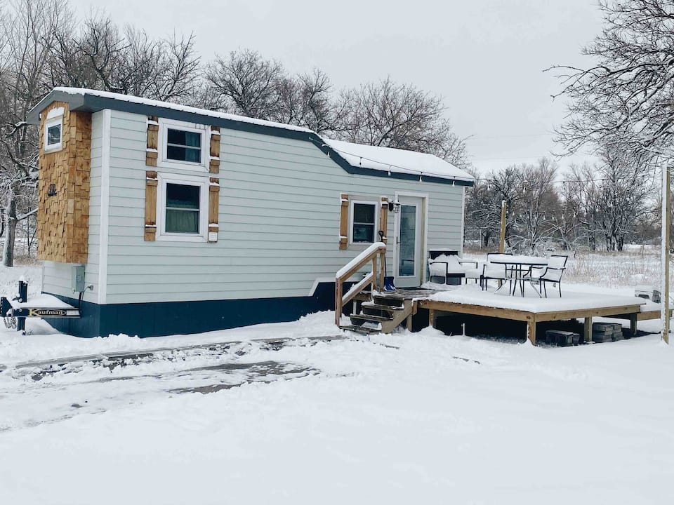 A tiny home covered in snow with a large front deck and patio set and natural wood details