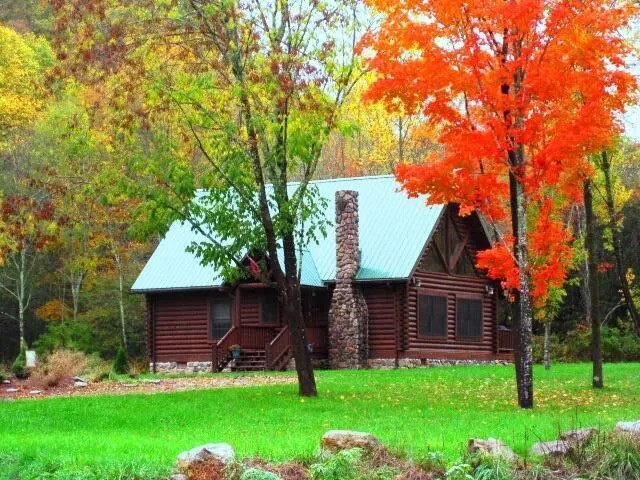 Brown log cabin with stairs and stone chimney surrounded by autumn leaves on trees.