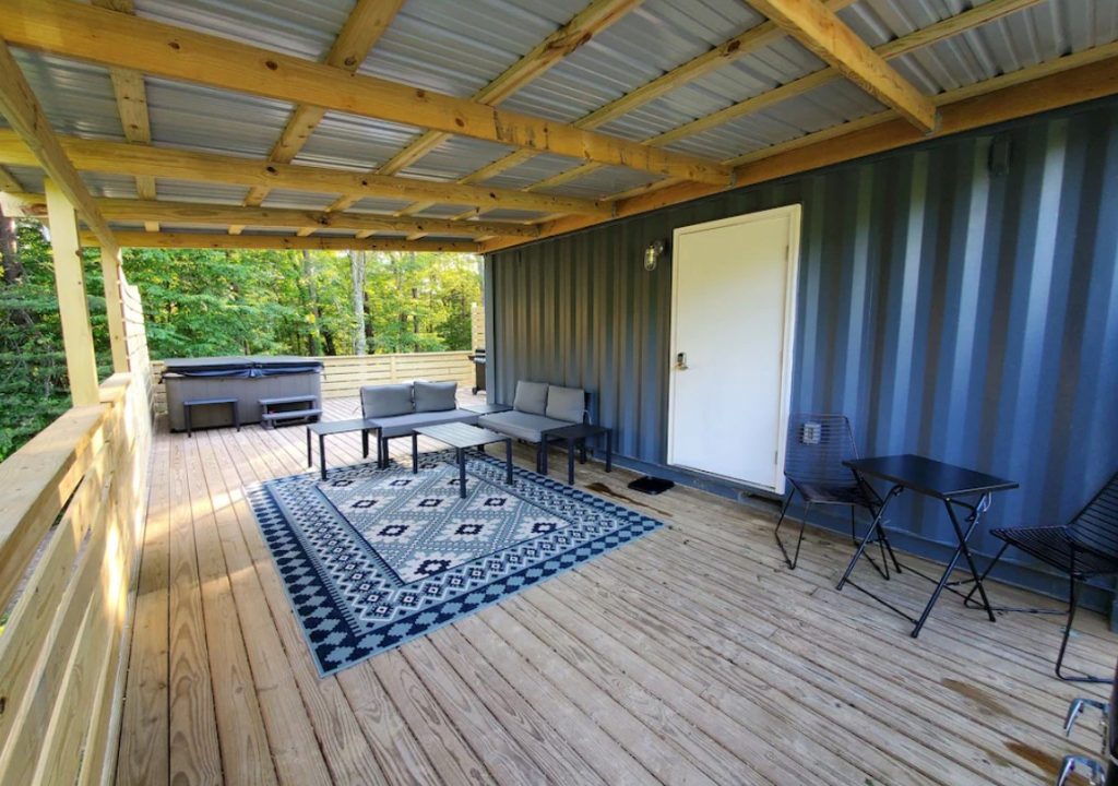 A large covered front porch on the side of a gray shipping container. There is black and grey patio seating, a rug that has a geometric design in shades of blue, and a hot tub. Behind the deck you can see trees. 