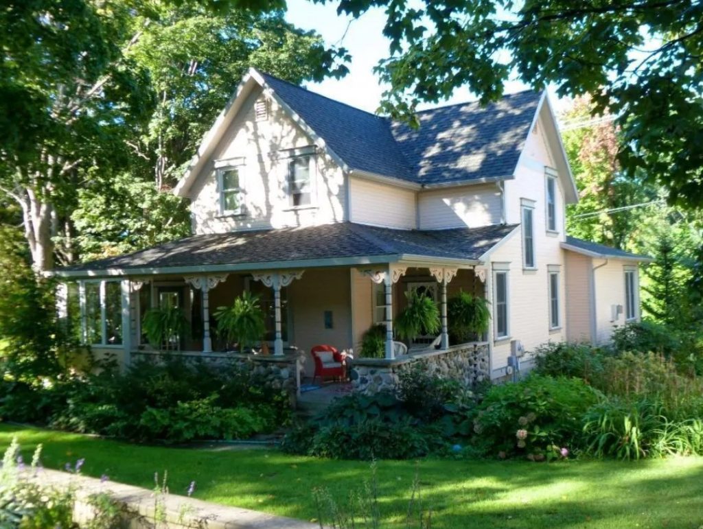The exterior of a historic home. It is a Victorian style home with a large front porch with a stone railing, decorative pieces on the columns, and ferns hanging from the ceiling of the porch. The house is painted a pale peachy pink and it is surrounded by shrubs and trees. 
