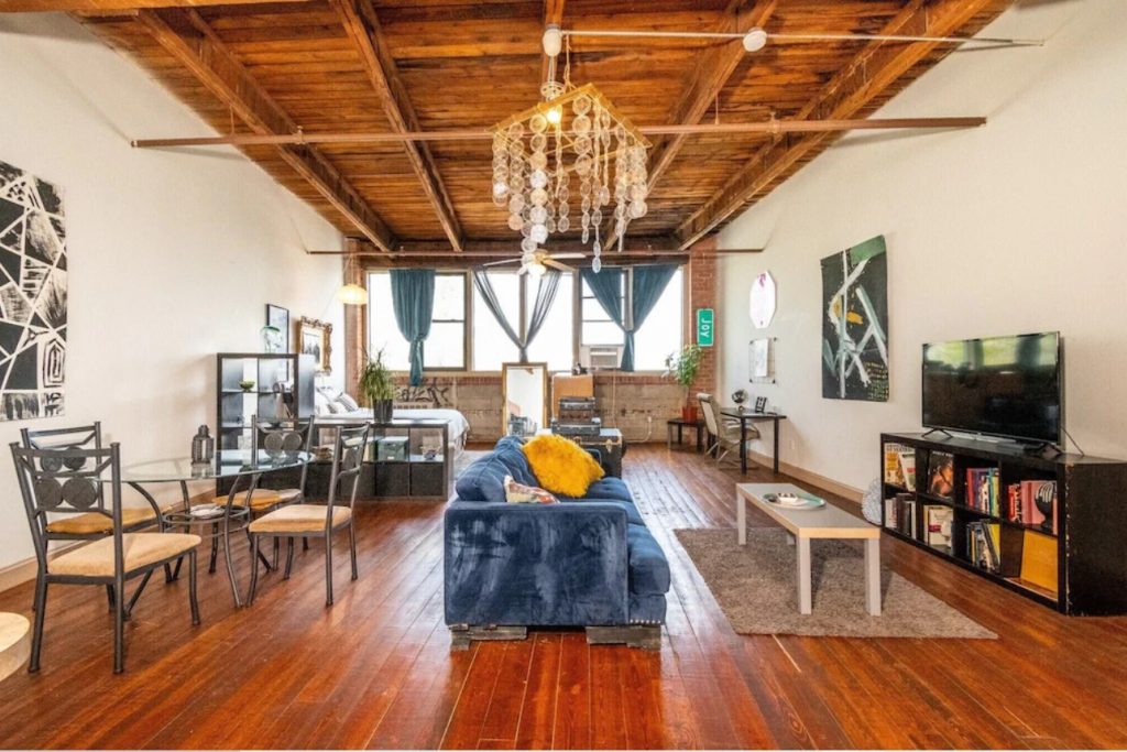 The inside of a loft in Detroit. It has hardwood floors, wood ceilings, a chandelier with discs, and large windows on the wall at the end of the loft. There is a velvet blue sofa, a dining set, a tv on a tv stand, and artwork on the walls. 