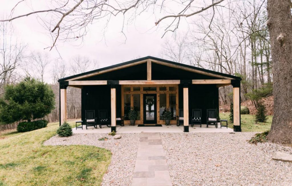 A large black cabin with a covered front porch and natural wood columns. It is on a grassy lawn and there is a pebbled area in front of the front porch. There are trees behind it with no leaves on them. One of the best cabins in Hocking Hills