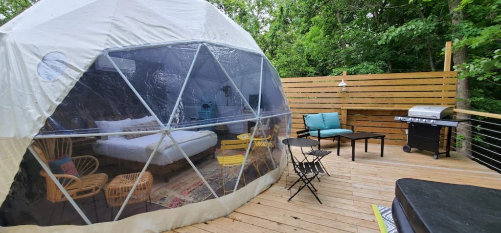 A white and see through dome on a large wooden platform in the woods. Through the clear areas of the dome you can see a bed and some chairs. On the platform there is some patio seating, a grill, and a hot tub. One of the best cabins in Hocking Hills