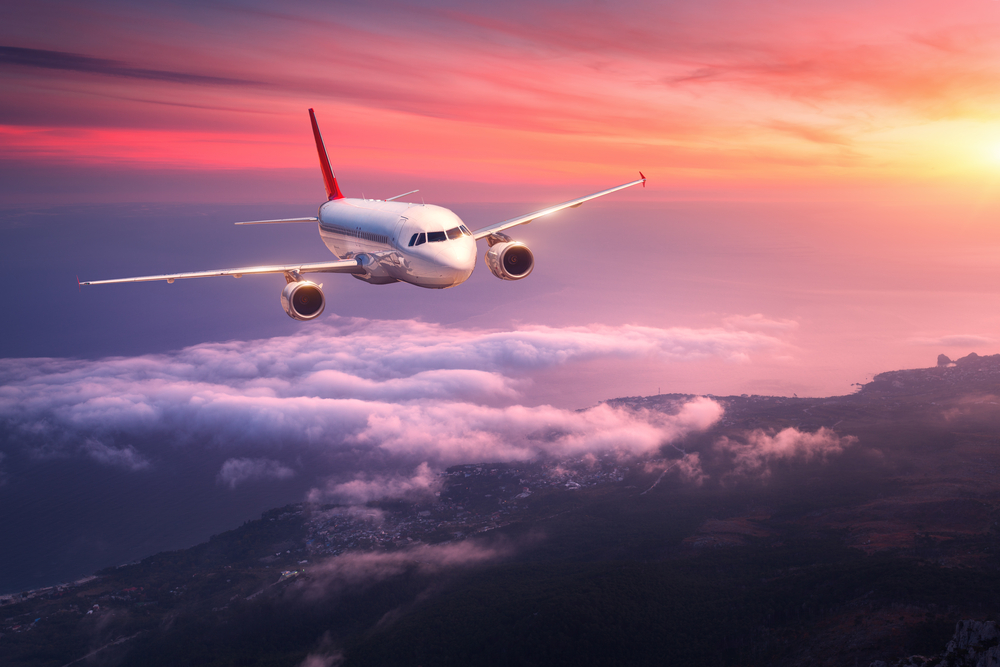 Passenger airline flying in red sky and brilliant sunset, over clouds, with sea below.