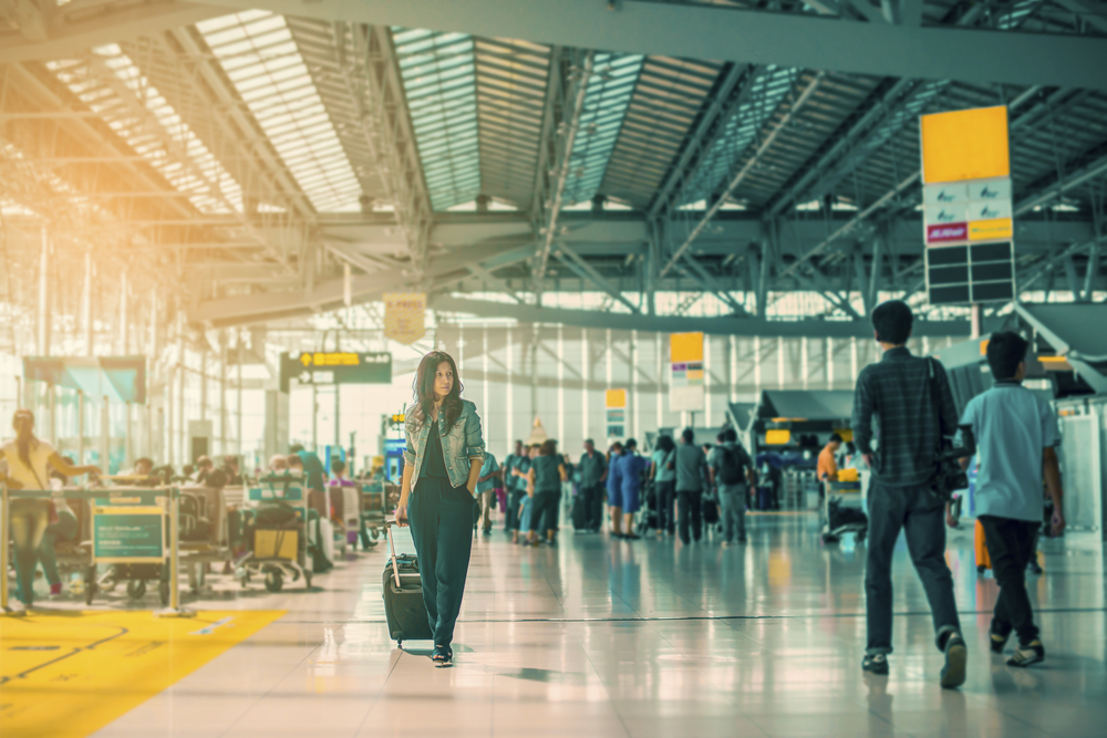 Passengers walk in large airport terminal. Female walking towards camera wheeling carry-on bag.