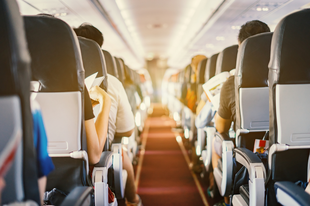 Interior of crowded airplane full of passengers on both sides of aisle with red rug in middle of photo.