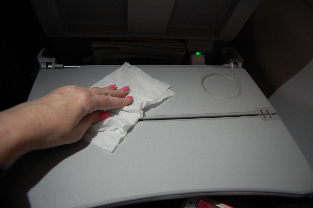 Female passenger with pink nail polish disinfects tray table on plane with wipe.